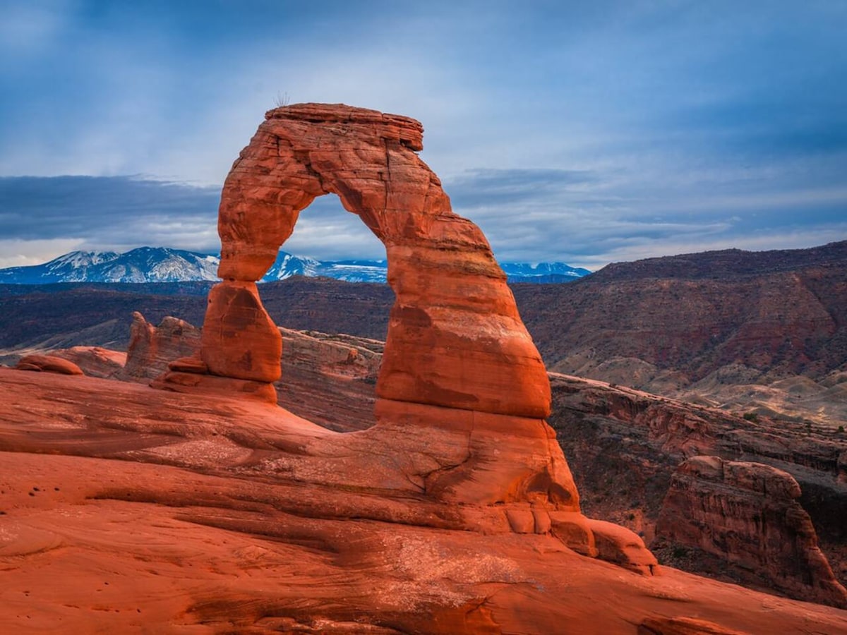 A prominent natural arch stands against a backdrop of rugged red rock formations. The arch features a smooth, rounded top with a wide opening below, framed by distant mountains under a cloudy sky. The surrounding terrain exhibits a blend of earthy tones and textures.