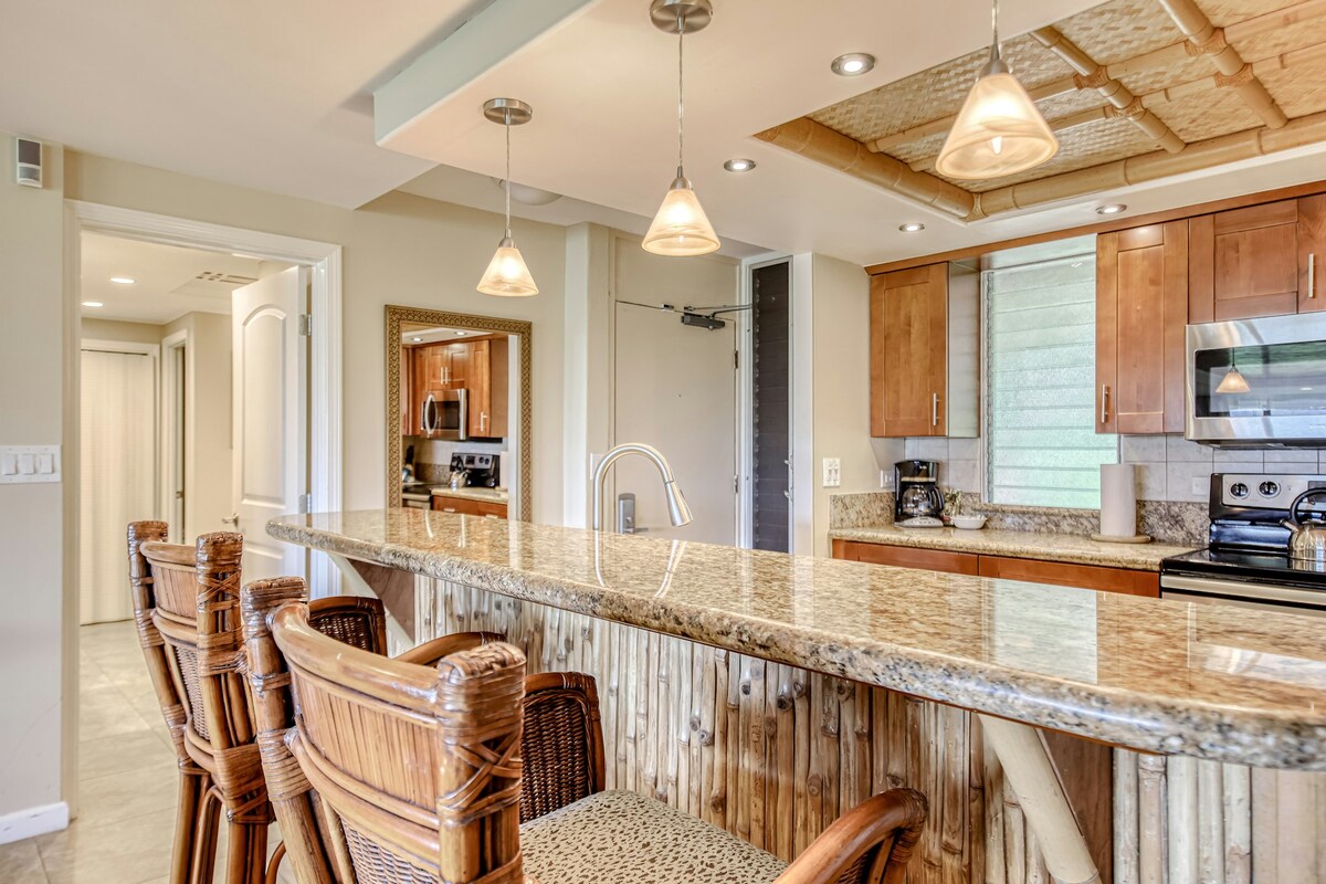A modern kitchen features a spacious granite countertop with four wicker bar stools positioned underneath. Natural light illuminates the room through a window, highlighting the bamboo ceiling accents. Full-size appliances and a clean, organized space can be seen in the background.