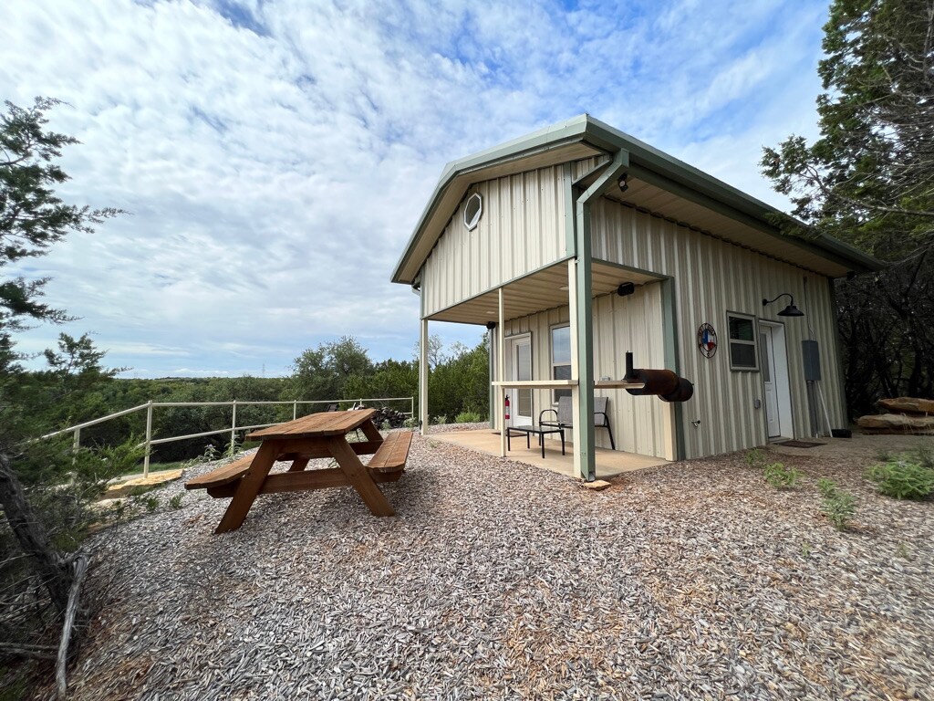 A cabin featuring a covered patio is shown, with a picnic table positioned on a gravel surface. The structure has a light-colored metal exterior and is surrounded by greenery, offering scenic views of the natural landscape.