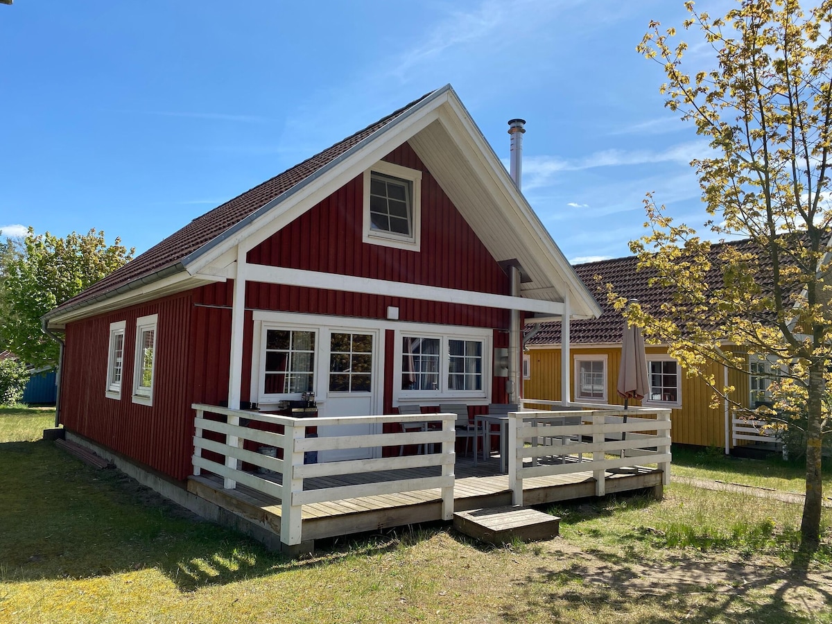 A charming red wooden house with a welcoming front porch is displayed, featuring white railings and outdoor seating. The gabled roof enhances the structure's character, while surrounding greenery offers a serene outdoor space. Nearby houses in varying colors complement the cheerful atmosphere.