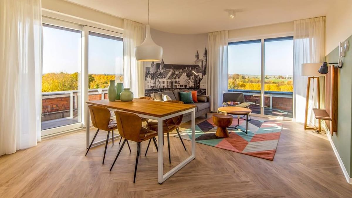 A spacious living area includes a dining table paired with four wooden chairs, and a comfortable seating arrangement with a large sofa facing large glass doors. Natural light floods the space, highlighting a colorful rug and a wall featuring a scenic mural. Scenic views are visible through the windows.