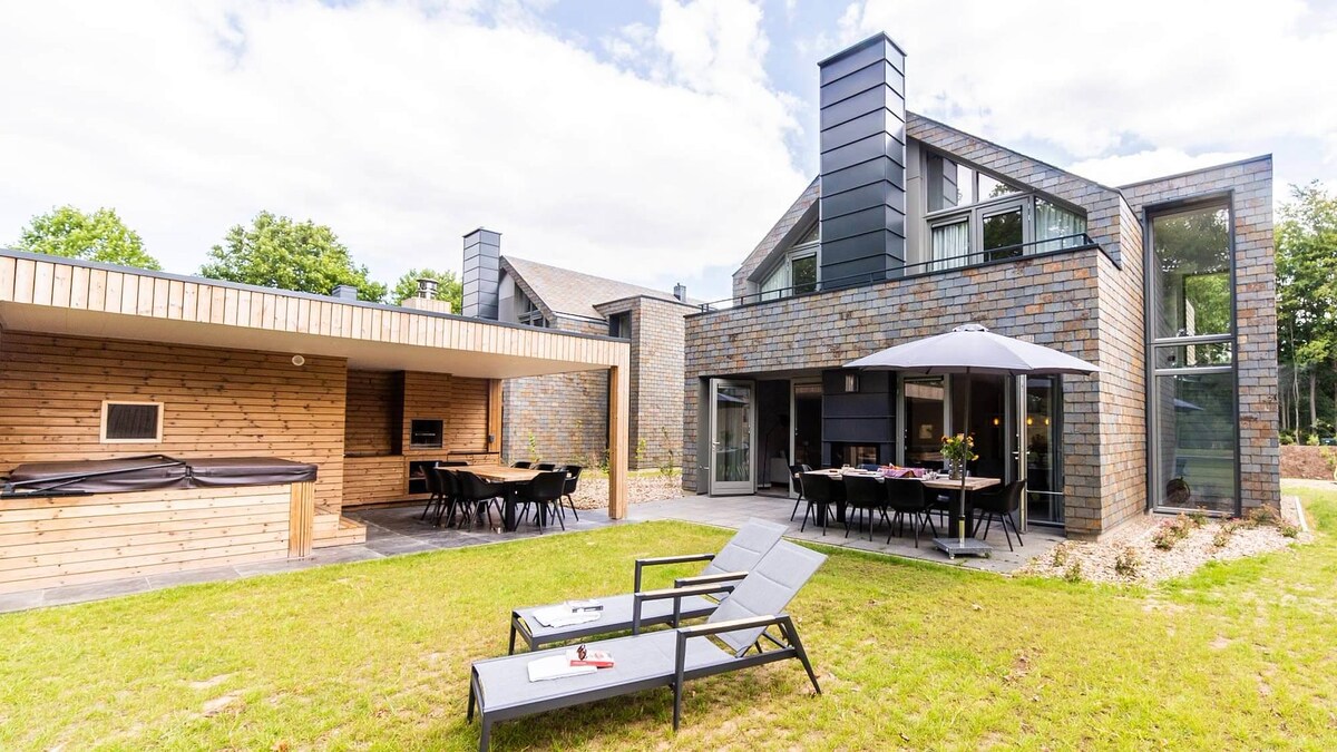 A modern villa with a stylish outdoor terrace and dining area is set in a landscaped garden. Two sun loungers are positioned on the grass, providing a relaxing space. An outdoor kitchen and barbecue area enhance the outdoor amenities, while large glass doors invite natural light.