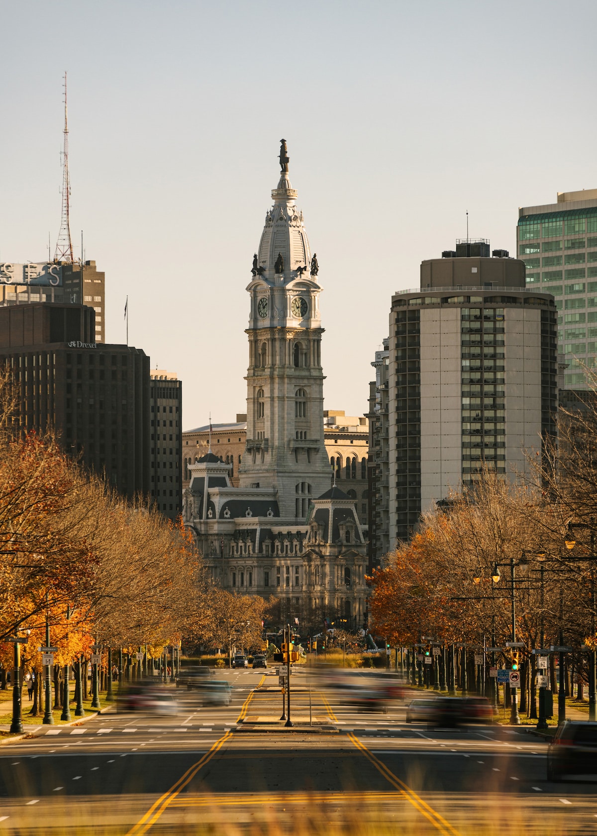 A prominent view of Philadelphia's City Hall is presented, featuring its iconic clock tower and intricate architectural details. Flanked by modern skyscrapers, the scene illustrates a tree-lined avenue with autumn foliage, capturing a blend of historic and contemporary urban landscapes.