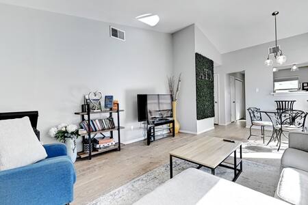 A bright living area features a cozy blue sofa and a coffee table positioned on a light-colored area rug. A small dining table with elegant chairs is visible, alongside a television mounted on the wall. Decorative plants add a touch of greenery.