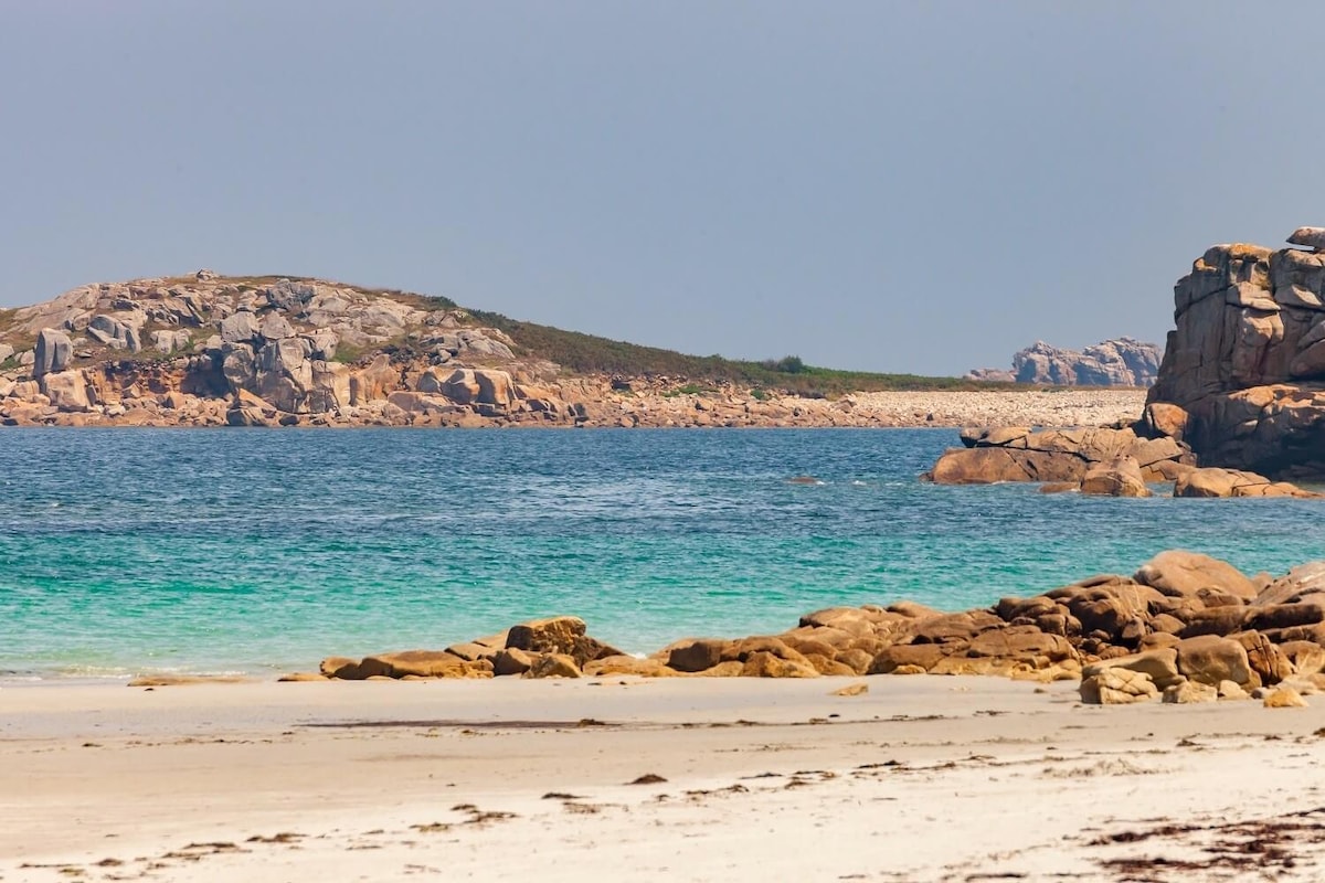 The image captures a scenic view of a tranquil beach with soft white sand and layered rock formations. Azure waters gently lap against the shore, transitioning into deeper shades near distant islands. The sky is clear, enhancing the bright colors of the landscape.