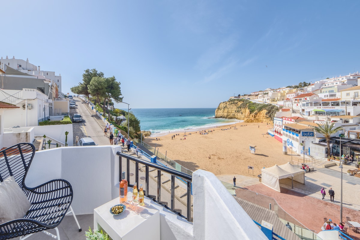 A balcony view overlooks a sandy beach framed by gentle waves and a clear blue sky. The beach is lined with a promenade, where visitors stroll. Coastal hills and white buildings provide a picturesque backdrop, enhancing the serene atmosphere.