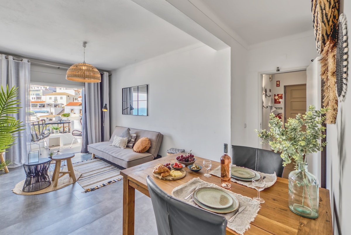 A modern living area is showcased with a cozy sofa and a dining table set for two. Natural light filters through the balcony doors, highlighting the tasteful décor. A vibrant plant adds a touch of greenery, complementing the airy space and inviting atmosphere.