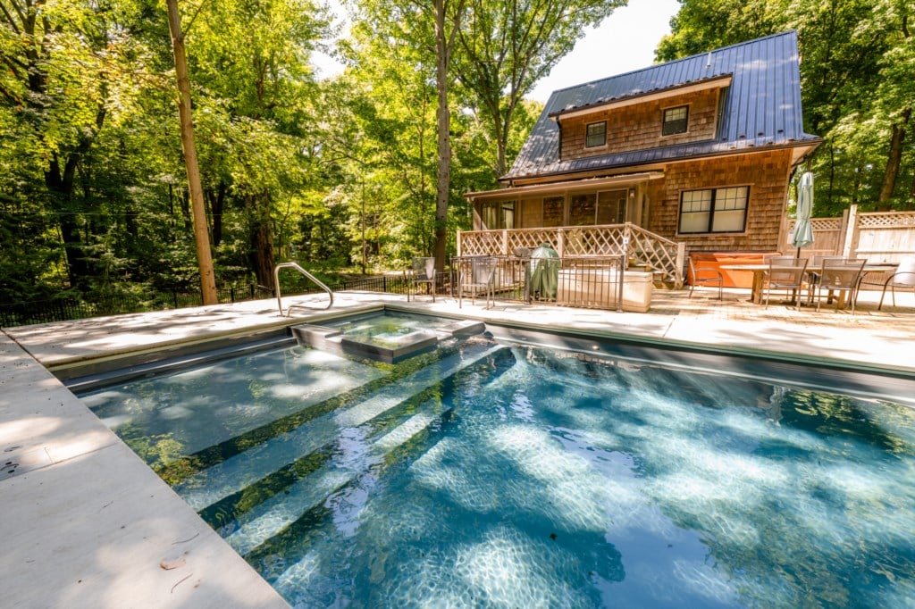 A private heated pool is surrounded by a spacious deck shaded by lush trees. A hot tub is positioned nearby on the deck, complementing a rustic cabin with a blue metal roof. Outdoor furniture provides a comfortable space for relaxation.