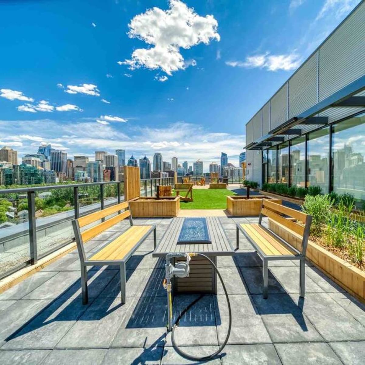 A spacious rooftop patio features wooden seating and a fire pit, framed by glass railing overlooking Calgary's skyline. Lush greenery and a communal barbecue area are also visible, creating an inviting space for socializing amidst the cityscape.
