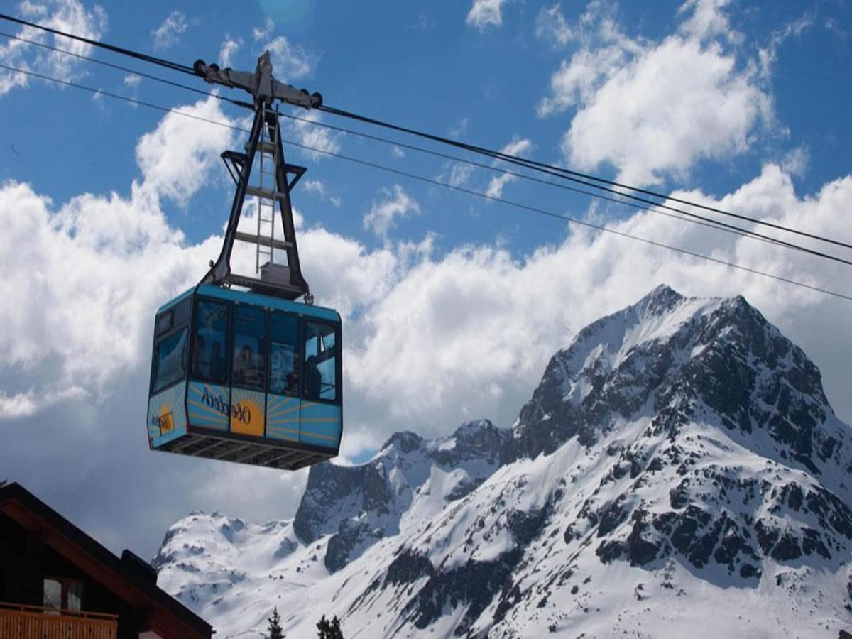 A cable car is seen traveling along its suspended track, set against a backdrop of snow-covered mountains. The structure is surrounded by a clear blue sky dotted with clouds, illustrating a scenic alpine environment.