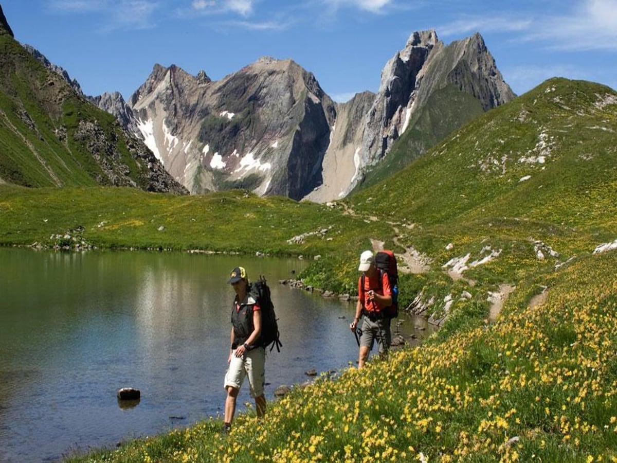 Two hikers are seen walking along the edge of a serene lake, surrounded by lush green meadows adorned with yellow wildflowers. Majestic mountains form a dramatic backdrop, showcasing rocky peaks and patches of snow under a clear blue sky.