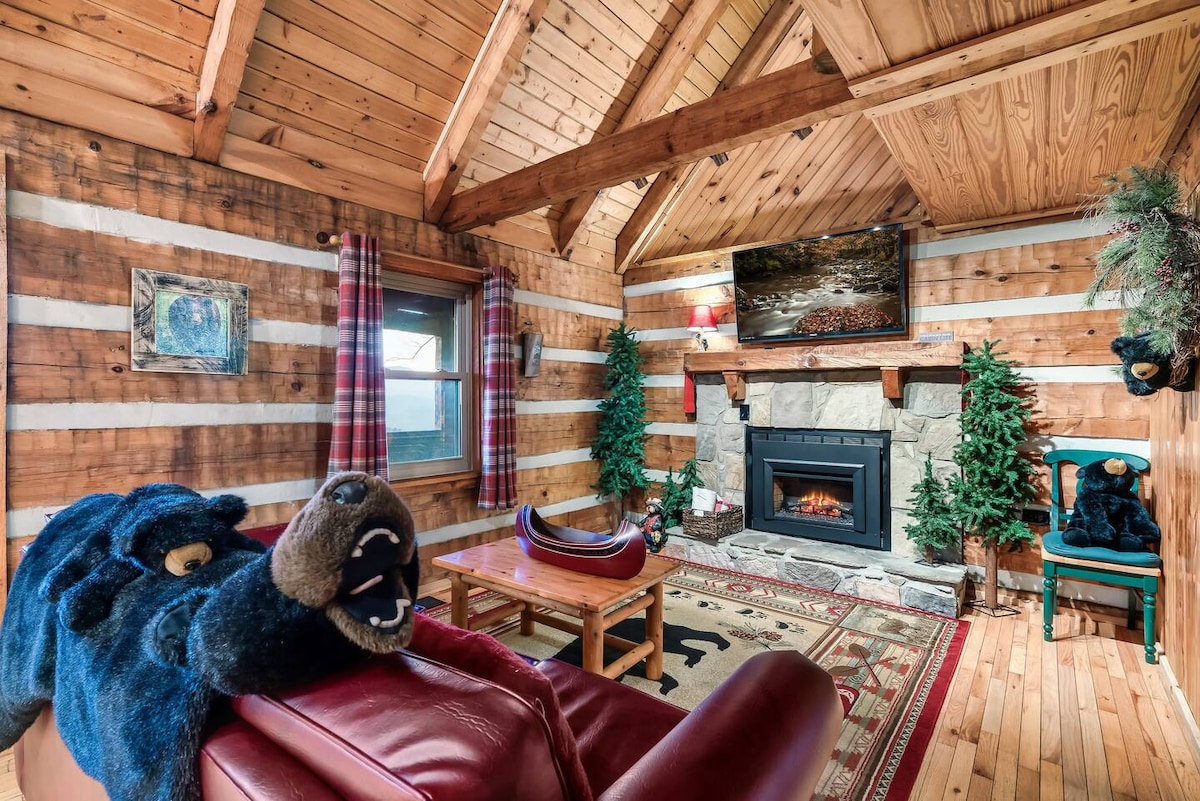The inviting living area features rustic wooden beams and log cabin walls. A plush red sofa is centrally positioned, adorned with a bear-themed throw. A stone fireplace adds warmth, with a mounted TV above, while green pines frame the space, enhancing the cozy atmosphere.