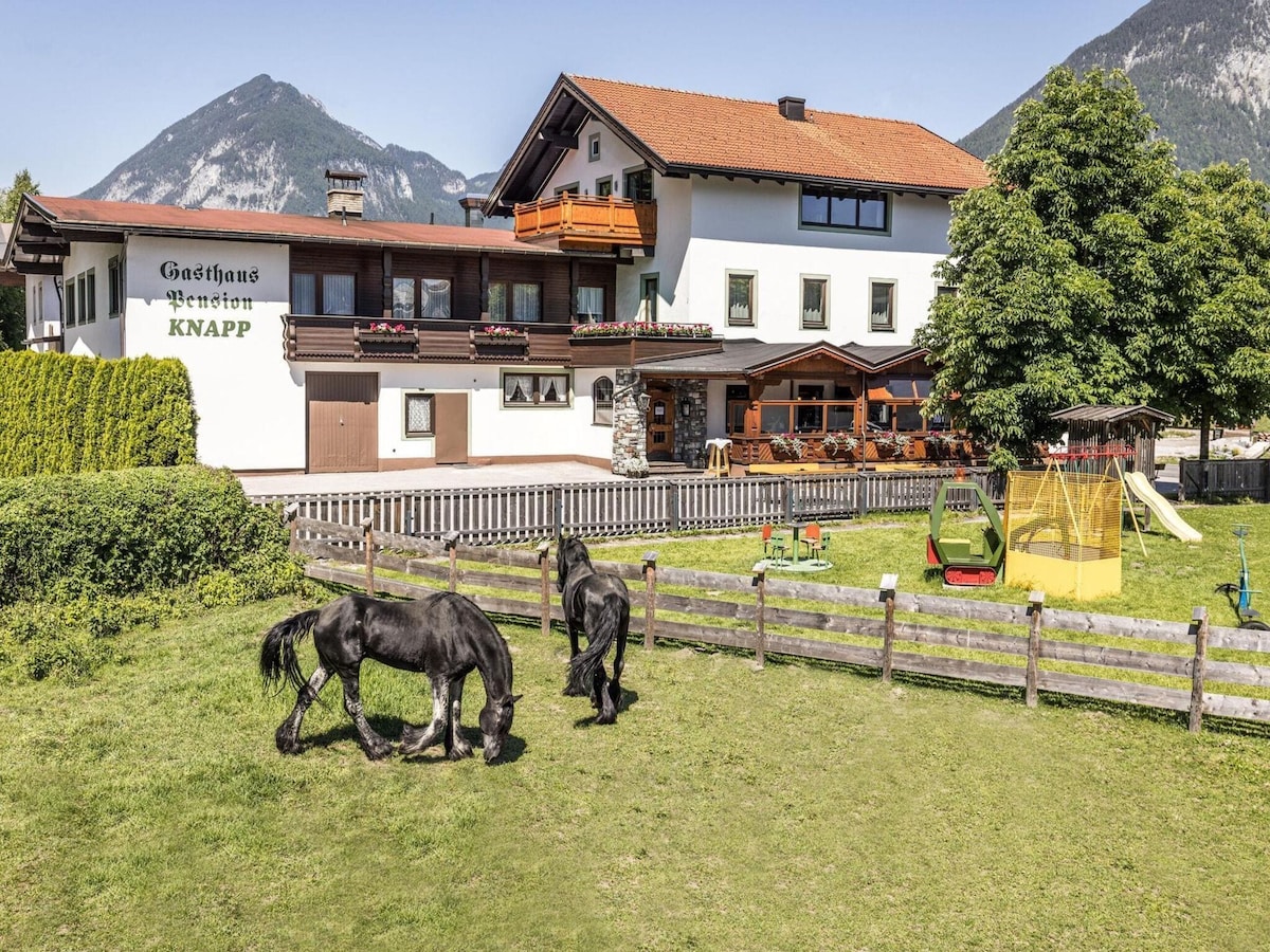 A spacious garden area features horses grazing peacefully on lush green grass, framed by a wooden fence. The multistructure building showcases balconies with vibrant orange accents and outdoor seating, with picturesque mountains in the background providing a natural setting.
