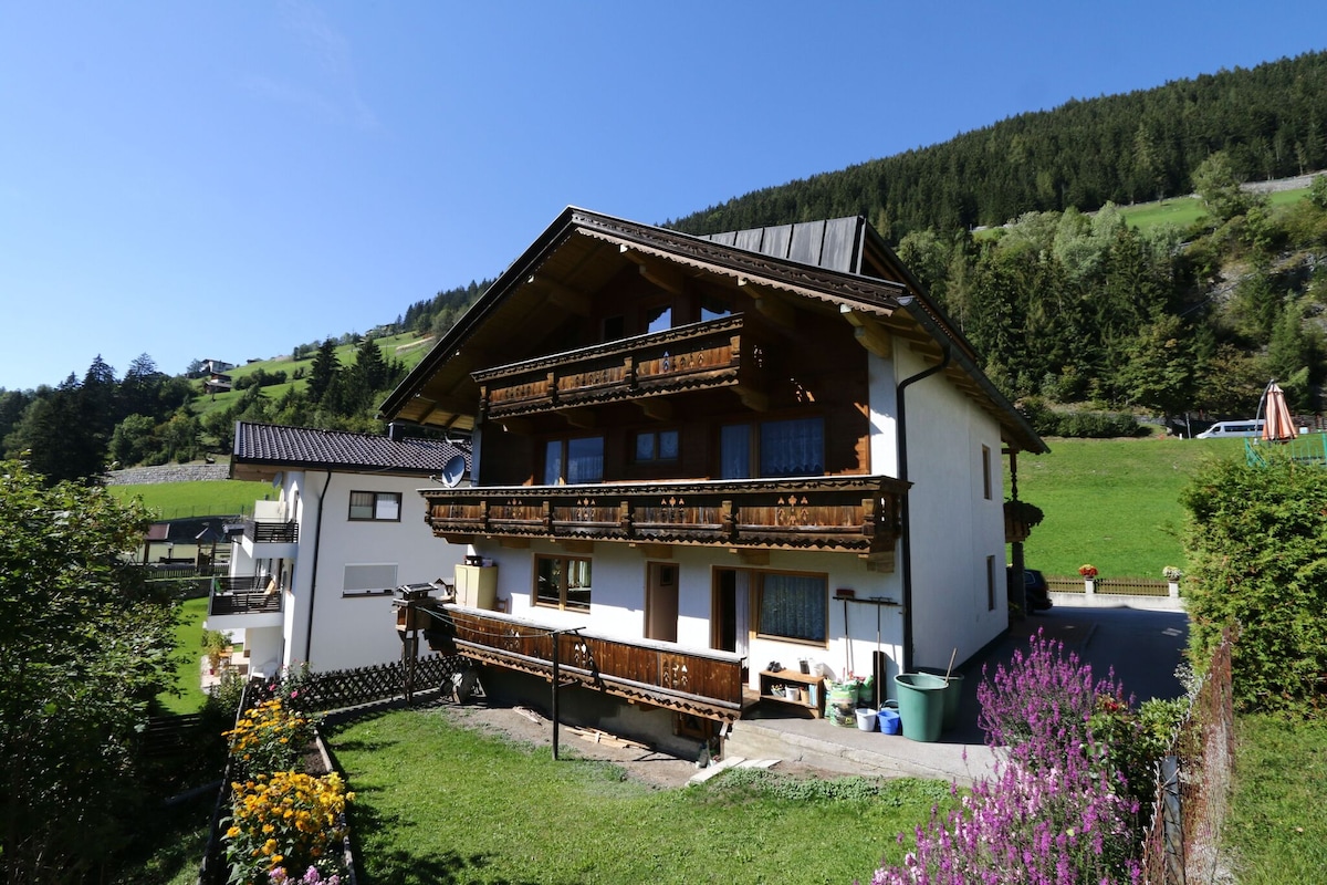 The exterior of the house is viewed from the ground, showcasing multiple balconies adorned with wooden railings. The building is surrounded by lush green grass and mountains in the background, creating a natural setting. Colorful flowers and greenery line the foreground.