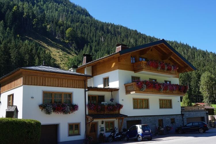 A charming multi-family building is set against a backdrop of lush green mountains. The exterior showcases wooden accents and vibrant flower boxes on balconies, adding a touch of natural beauty. A driveway leads to parking areas, offering easy access to the entrance.