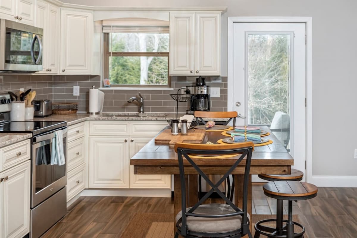 A modern kitchen features cream-colored cabinetry and stainless steel appliances, along with a wooden dining table set for four. Natural light enters through a large window, revealing views of the surrounding greenery. A doorway leads to a patio area.