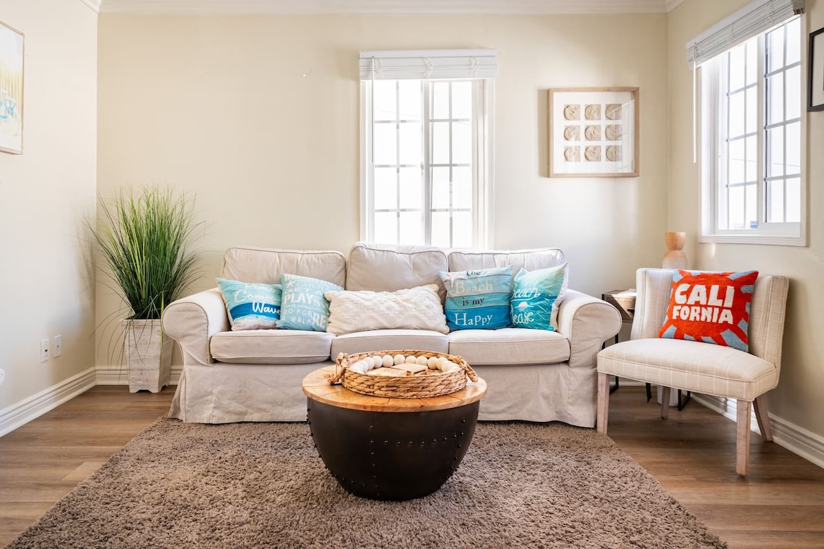 A light-filled living area features a comfortable sofa adorned with thematic cushions. A round coffee table sits at the center, surrounded by a plush rug. Sunlight streams through the windows, highlighting the neutral wall tones and the greenery positioned nearby.