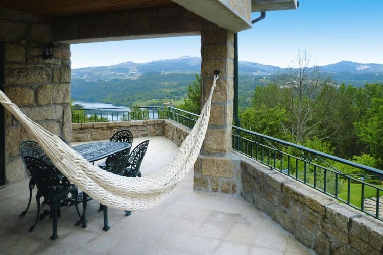 A stone terrace is shown with a large outdoor table and chairs, inviting for meals or relaxation. A hammock is displayed nearby, offering a comfortable spot to enjoy the scenic view of the TÂmega reservoir and surrounding greenery.