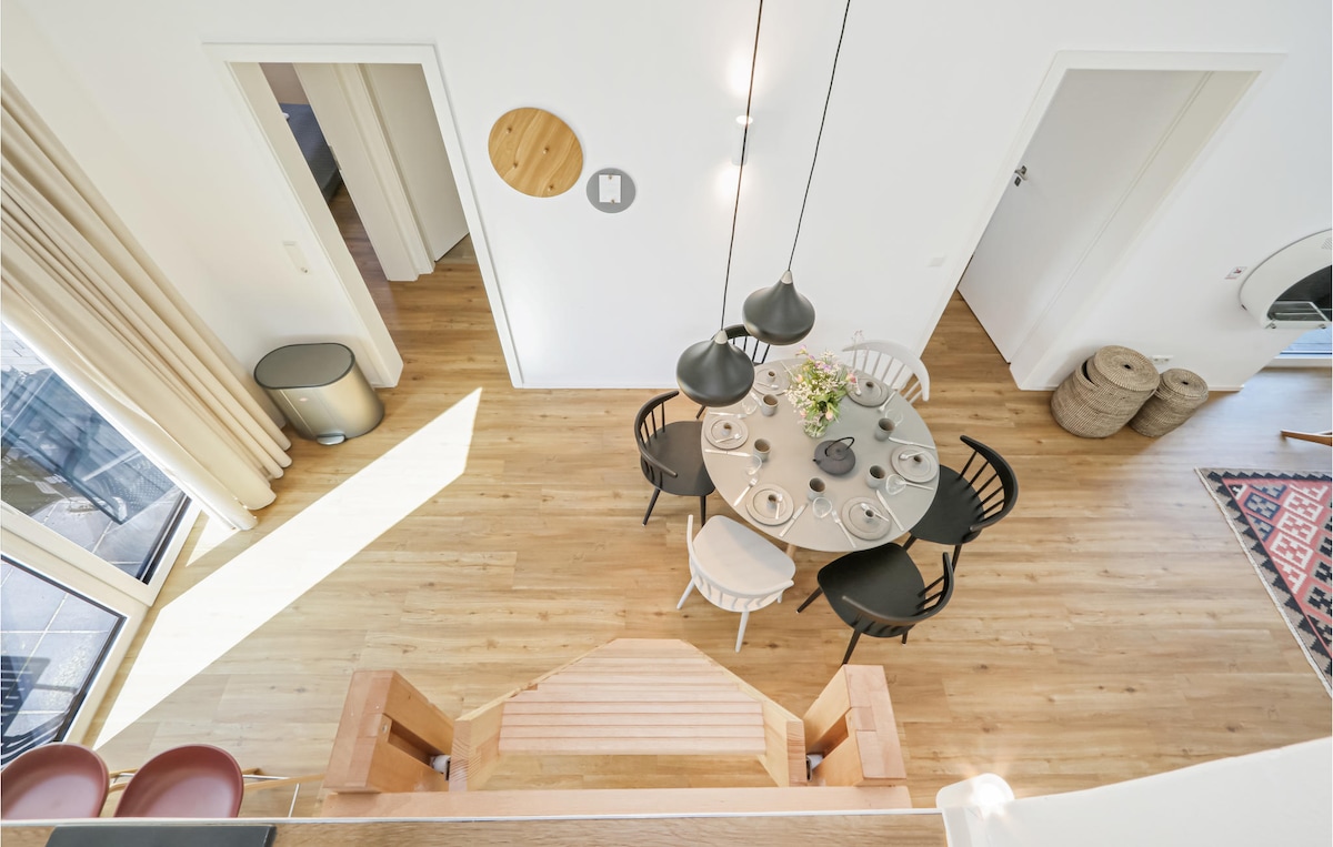 An overhead view of a dining area features a round table with six chairs in contrasting colors. Soft lighting is provided by three pendant lamps above the table. Light wood flooring is visible throughout the space, complemented by neutral-colored walls and large windows allowing natural light.