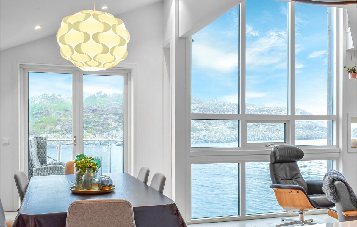 A dining area is presented with a large table surrounded by several chairs. A modern light fixture hangs overhead. Expansive windows offer a clear view of the sea and surrounding landscape, creating a bright and airy atmosphere.