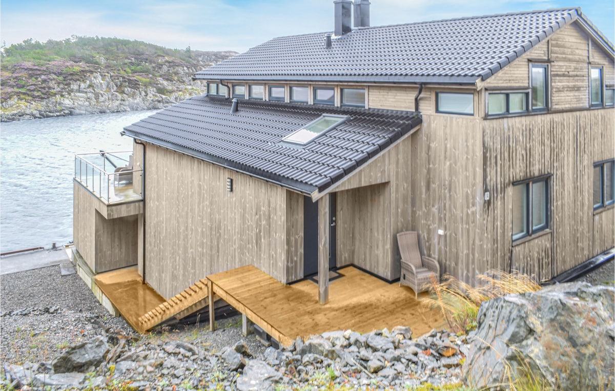 A large, modern vacation home featuring a wooden exterior and multiple windows. A wooden deck leads to an entryway, complemented by a comfortable chair. The sea is visible nearby, enhancing the distinctive coastal setting.