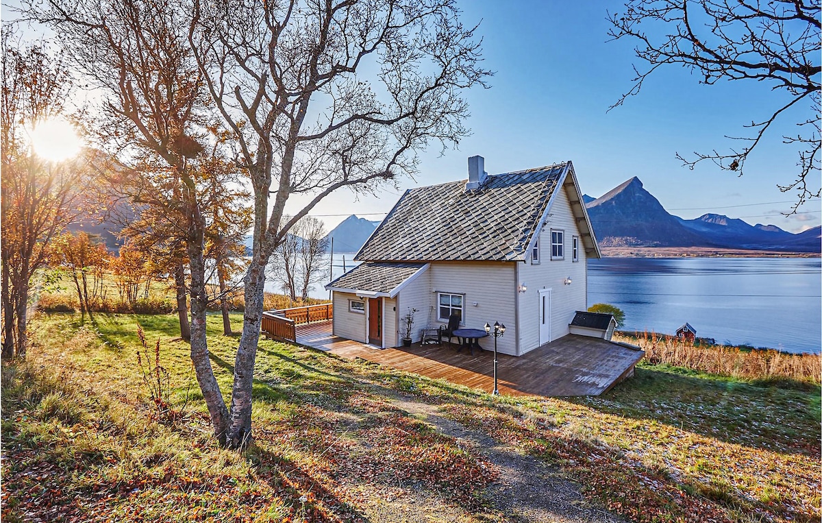A charming cottage is situated by a calm body of water, framed by mountains in the background. The wooden deck extends from the side, while trees and grasses surround the property. Early sunlight casts a warm glow over the scene, enhancing the natural beauty.