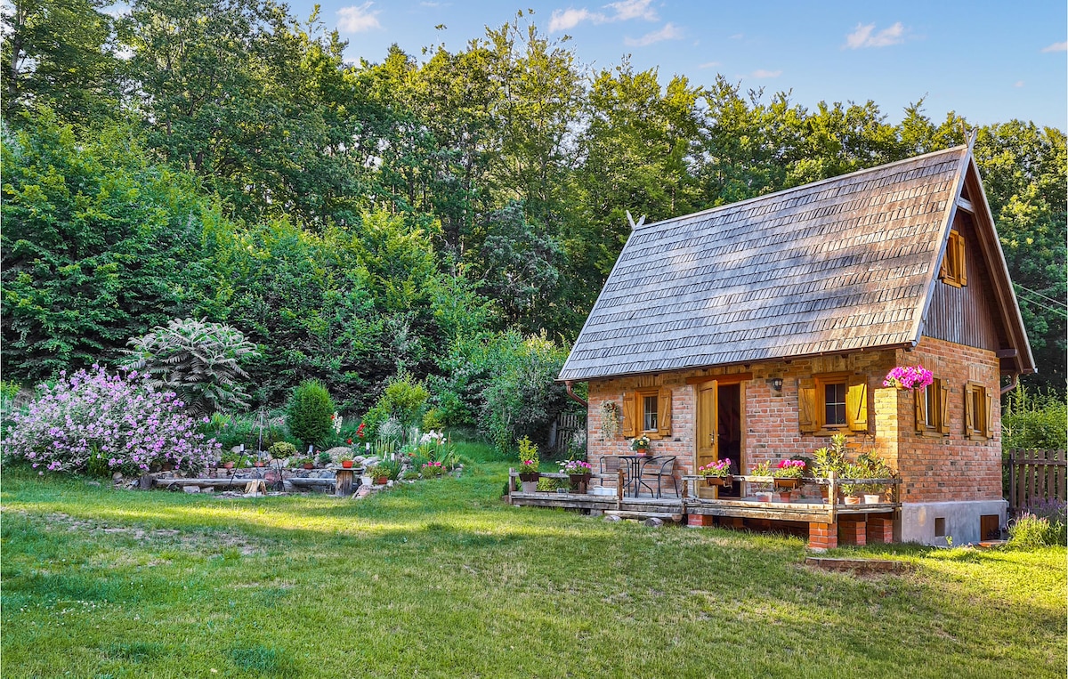 A charming holiday home is nestled in a lush green landscape, surrounded by trees and vibrant flower beds. The rustic structure features a sloped roof and multiple windows, while a large grassy area invites relaxation. A small patio with seating enhances outdoor enjoyment.