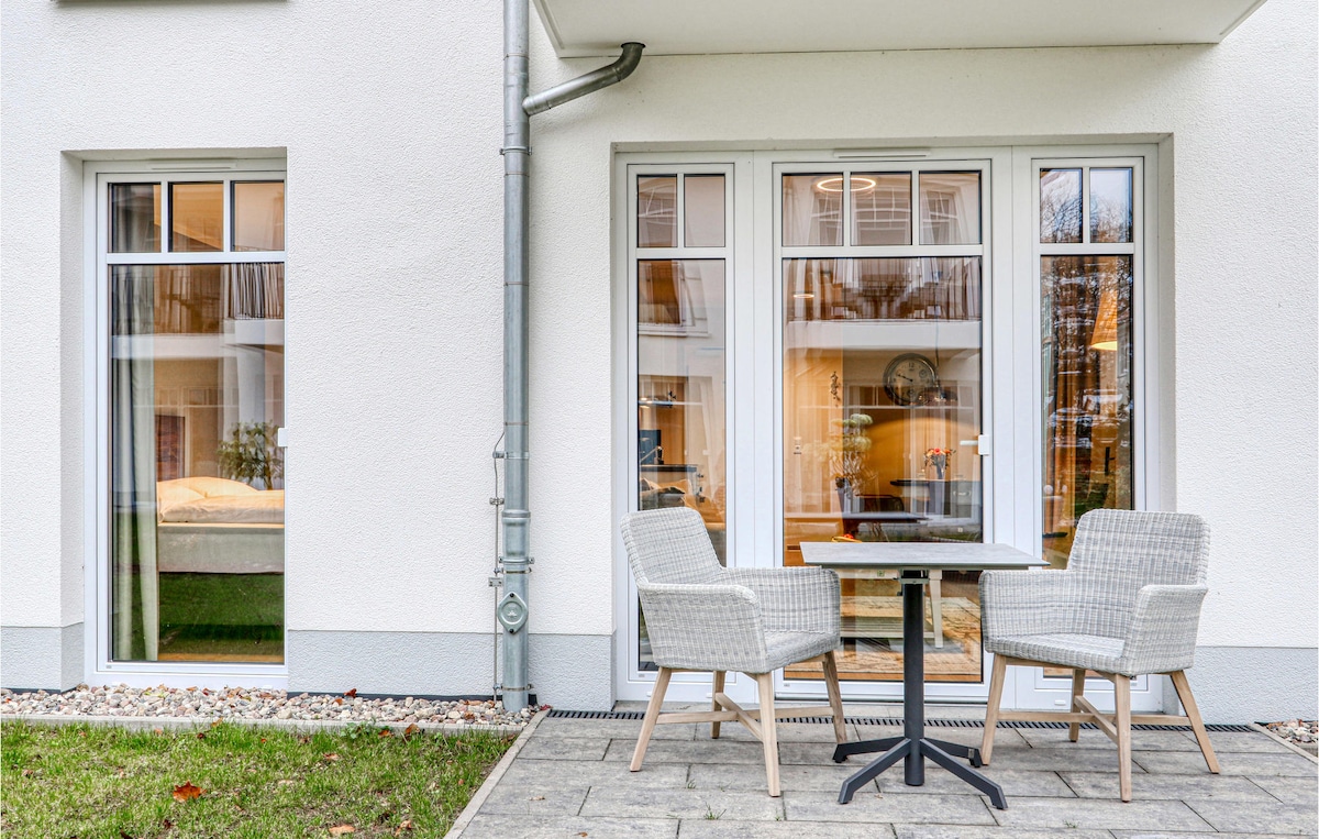 A terrace area features a small round table paired with two light-colored wicker chairs. Large glass windows provide natural light and a view of the interior. The surrounding space includes well-maintained greenery and attractive stone paving.