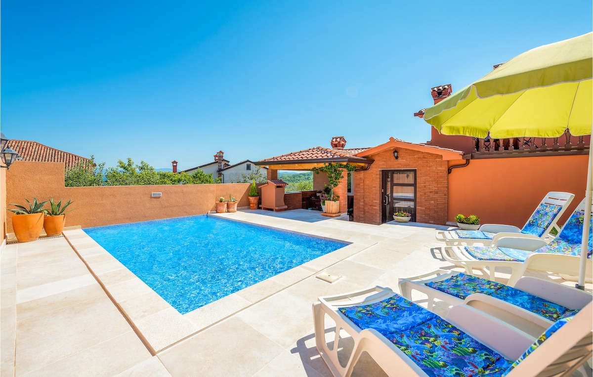 A spacious outdoor area features a sparkling blue pool surrounded by tan stone tiles. Colorful sun loungers are arranged nearby, complemented by a large green umbrella. A charming brick structure serves as an outdoor kitchen, with potted plants adding greenery to the space.