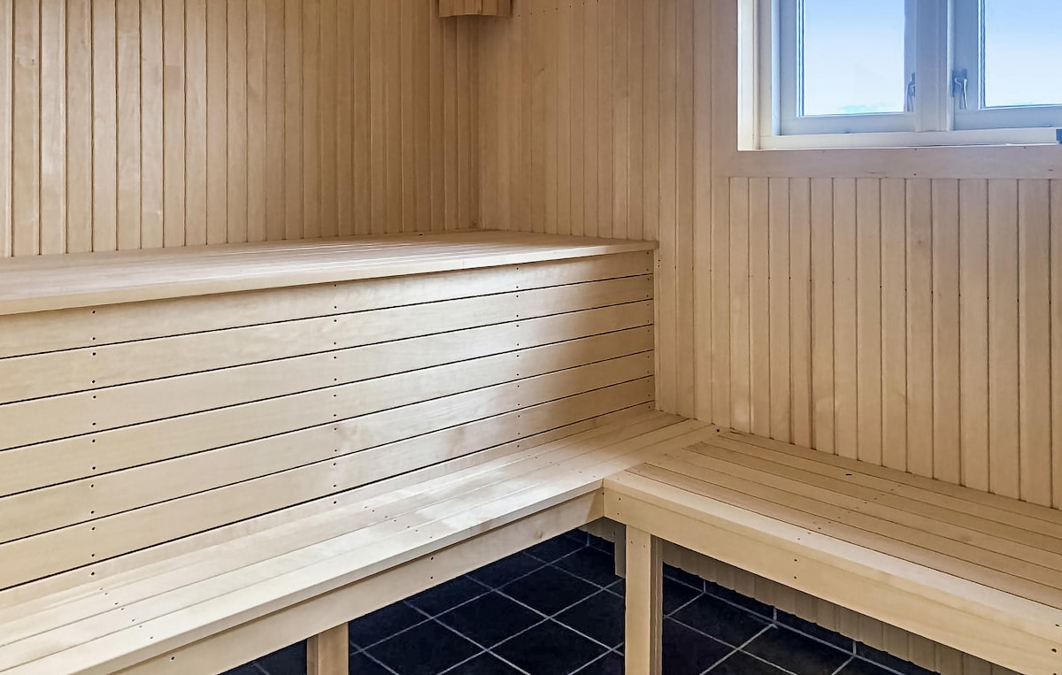 A wooden sauna interior features smooth, light-toned wooden benches and paneling, creating a serene environment. Natural light filters in through a window, illuminating the space. The floor is finished with dark tiles, providing a contrasting base to the warm wooden elements.