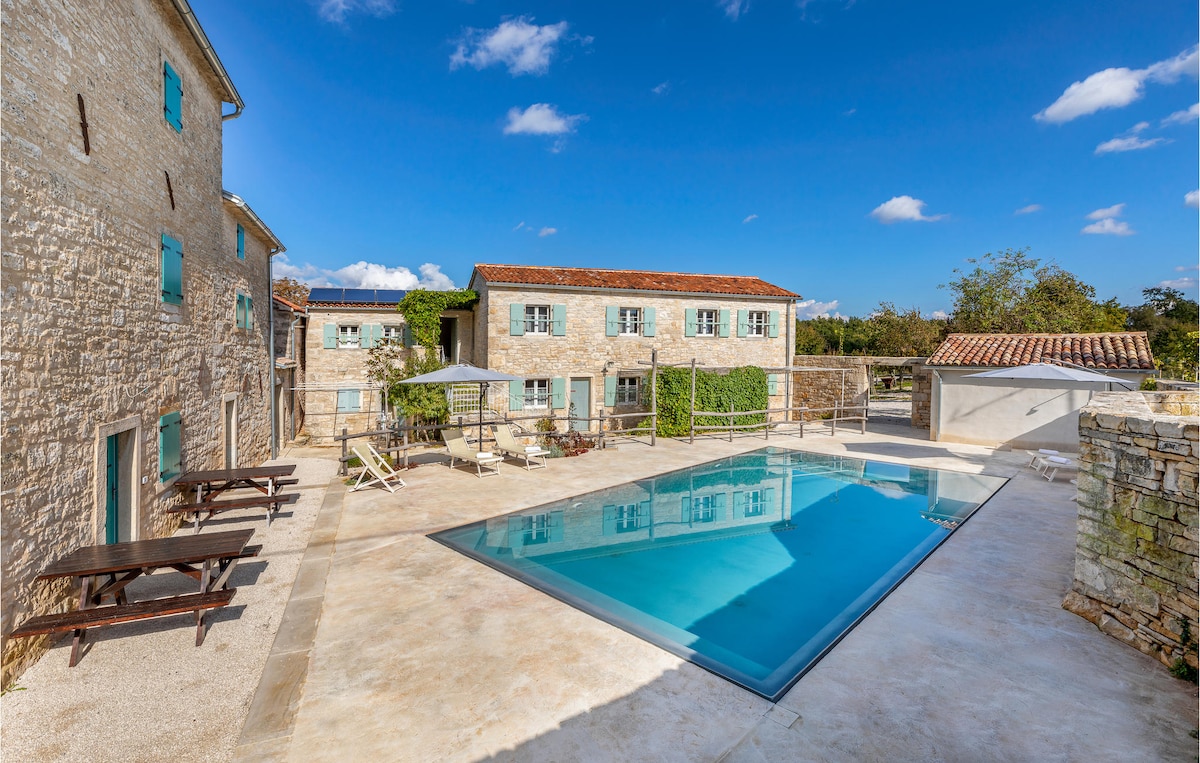 A shared outdoor pool is surrounded by a stone patio featuring sun loungers and shaded seating areas. The well-maintained buildings are characterized by charming stone walls and blue window shutters under a clear blue sky, creating a serene and inviting atmosphere.