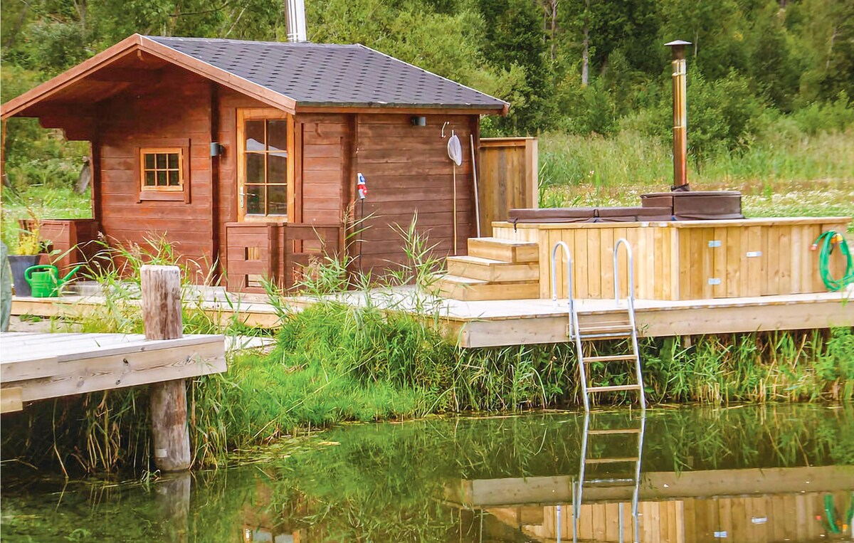 A wooden structure with a sloped roof is positioned near a tranquil water's edge, surrounded by lush greenery. A wooden deck with a stairway leads into the water, and a wood-fired hot tub is located on the deck, offering scenic views of the natural surroundings.