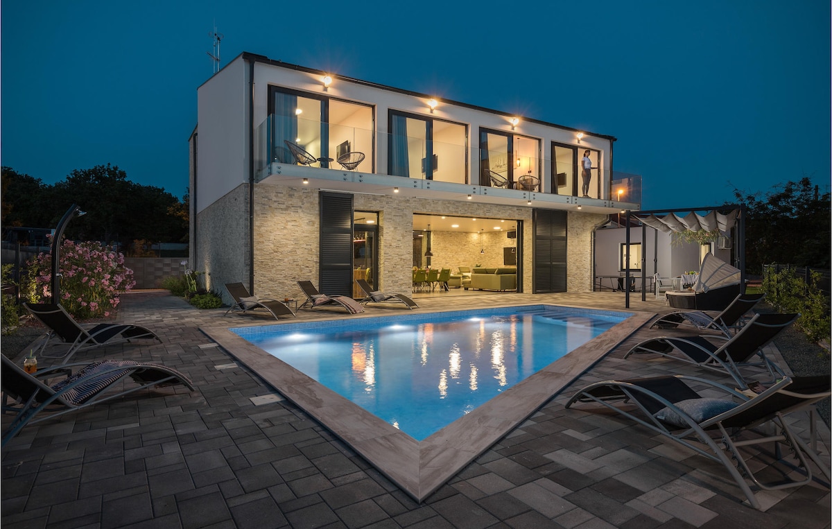A modern vacation home features a spacious outdoor area highlighted by a private swimming pool, illuminated against the evening sky. The two-story building has large glass doors revealing an open-plan living space, with comfortable loungers arranged neatly around the pool.