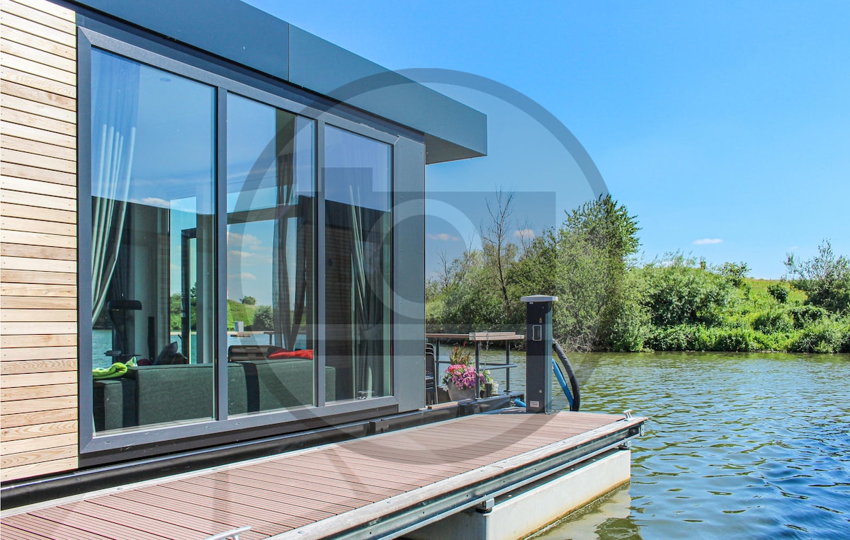 Large glass doors provide an unobstructed view of the serene water and surrounding greenery. The entrance features a wooden deck leading to the houseboat, with a subtle floral arrangement nearby. Sunlight reflects off the water, enhancing the natural setting.