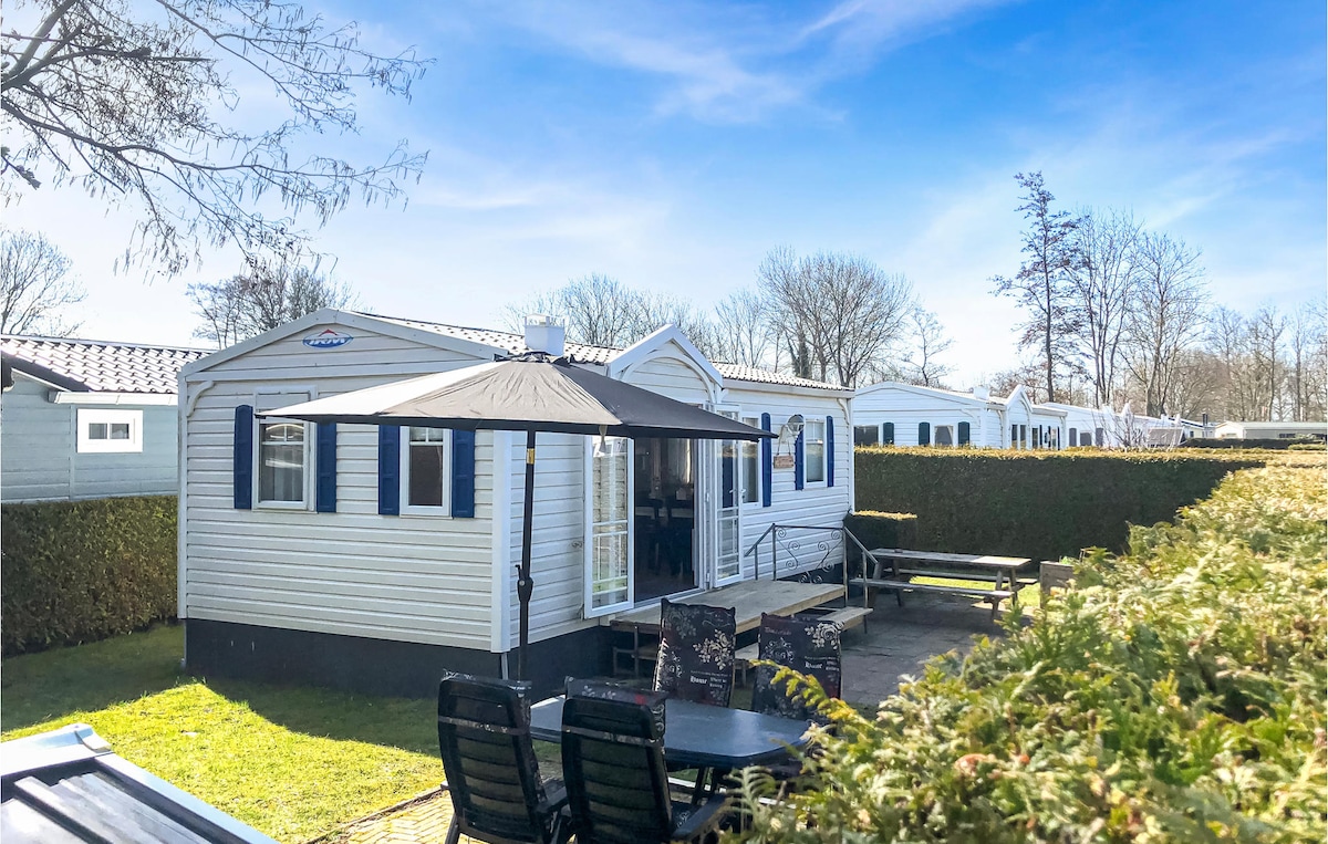 A charming cottage is surrounded by neatly trimmed hedges, offering a spacious outdoor area with a large umbrella and a set of black patio chairs. The light-colored exterior of the cottage features blue shutters, and nearby trees provide a mix of shade and sunlight.