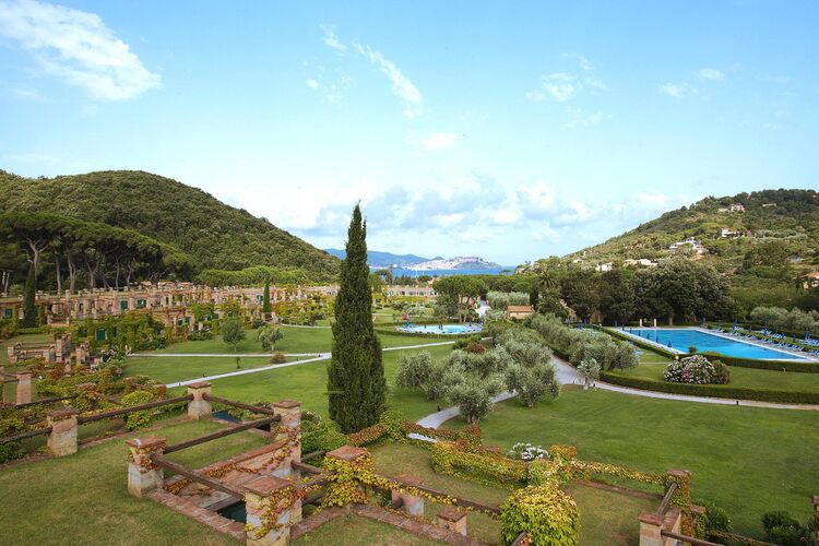 A landscaped garden is visible, featuring manicured lawns, ornamental trees, and flowering shrubs. Multiple swimming pools are situated amidst the greenery. Rolling hills create a serene backdrop, while a clear blue sky complements the peaceful setting.