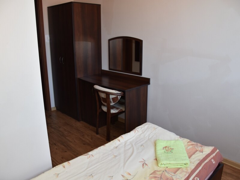 A simple guest room features a spacious bed with light bedding. A wooden dresser with a mirror is positioned beside a modest desk and chair. Warm wooden flooring complements the neutral wall color, creating a calm and functional environment.