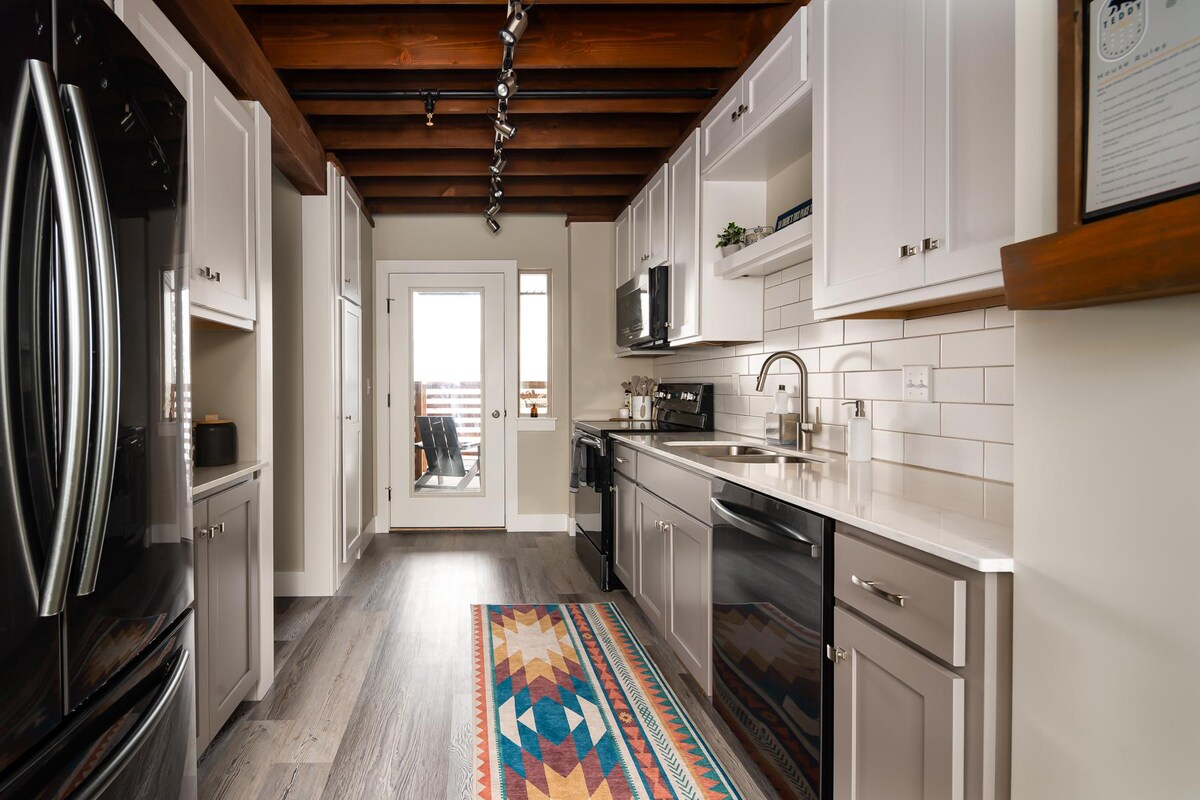 A modern kitchen is depicted, featuring sleek cabinetry, stainless steel appliances, and solid surface countertops. A colorful rug adds visual interest to the floor. Natural light is welcomed through glass doors that lead to a small deck, enhancing the kitchen's bright atmosphere.