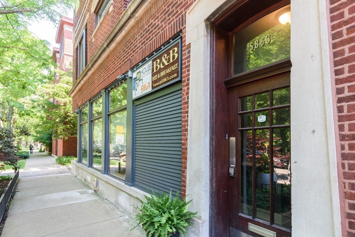 The exterior of the building features a classic brick facade complemented by large windows that allow natural light into the interior. Greenery is seen along the pathway, enhancing the inviting entrance with a wooden door and a visible signage for the bed and breakfast.