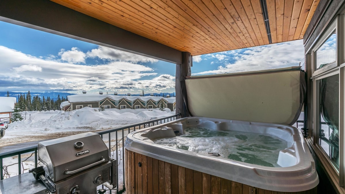 A private hot tub is situated on a covered patio, offering stunning views of the surrounding snowy landscape and distant mountains. A stainless steel BBQ is placed nearby, enhancing the outdoor space for gatherings or meals.