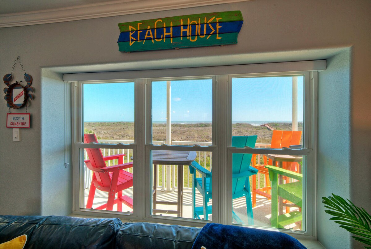 A cozy living area is shown with a blue sofa facing large windows. Brightly colored chairs on the private balcony offer scenic views of the coastal landscape. A wooden sign reading 'BEACH HOUSE' adds a cheerful touch above the window.