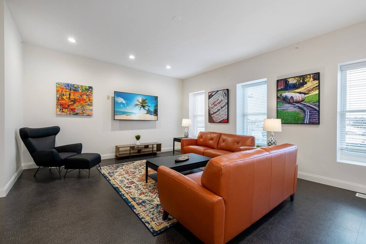 A modern living room features a sleek leather sofa and a stylish armchair. Bright artwork adorns the walls, while a large television is mounted for entertainment. Natural light enters through two windows, and a decorative rug adds comfort to the floor.