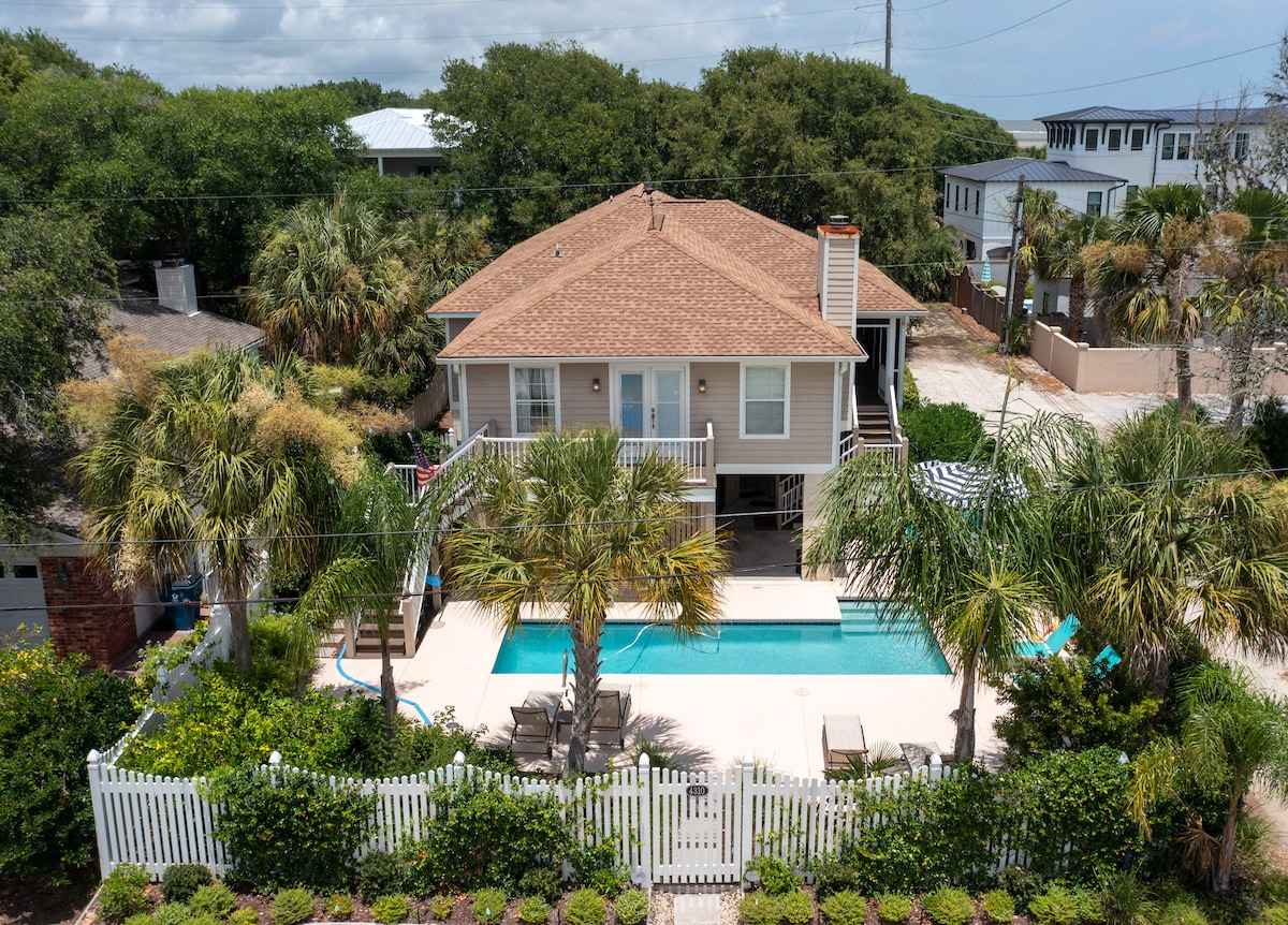 An aerial view of the property showcases a one-level home surrounded by lush greenery. A pool is visible in the backyard, complemented by a spacious patio area. Palm trees are strategically placed, while a white fence delineates the outdoor space.