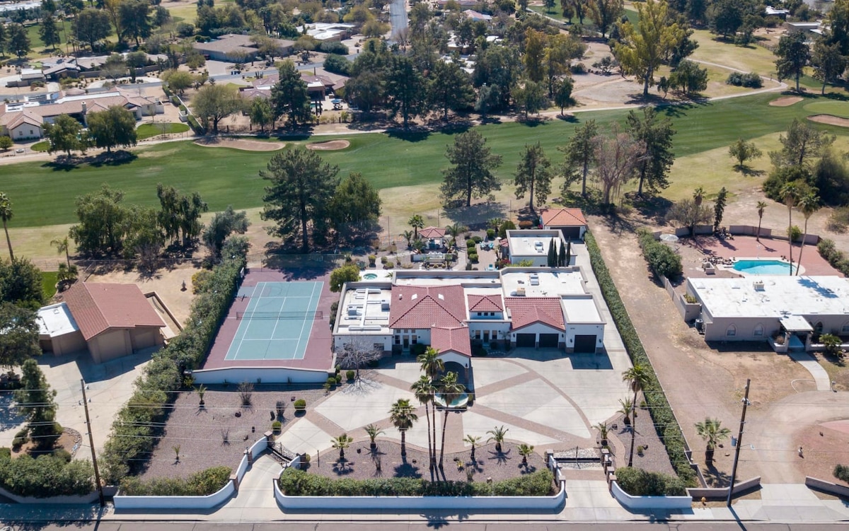 An overhead view captures the spacious property layout, featuring a tennis court alongside a well-manicured area with palm trees. The main building is complemented by separate structures, with a clear view of the golf course in the background.