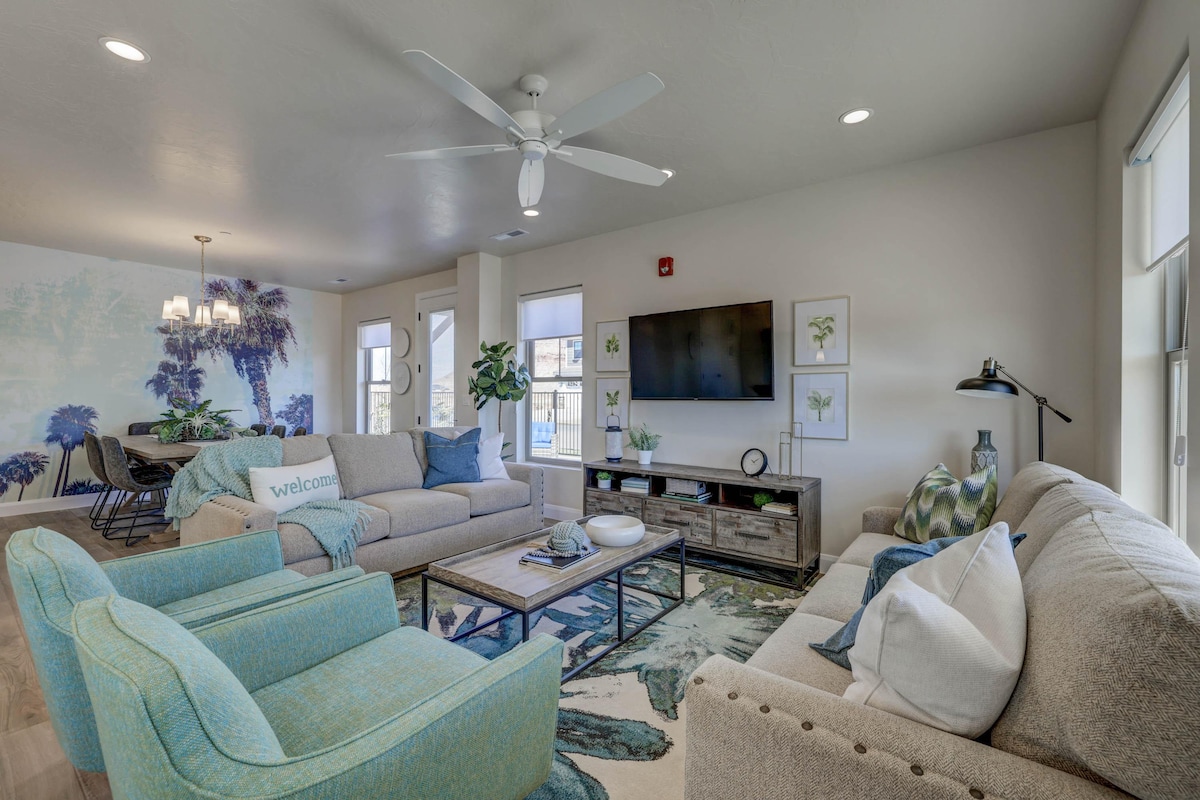 An airy great room features light-colored furnishings with a mix of seating options. A large wall mural depicting palm trees adds a tropical touch. A neutral area rug anchors the space, while a television is mounted above a sleek console table.