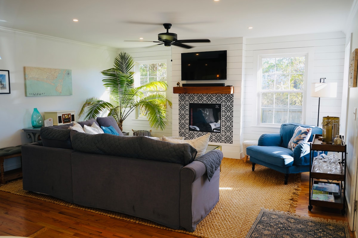 A spacious living area features a large sectional sofa with soft pillows, complemented by an accent chair. A modern fireplace is surrounded by intricate tile work, and a flat-screen TV hangs above. Large windows allow natural light to fill the room, with greenery adding a touch of nature.
