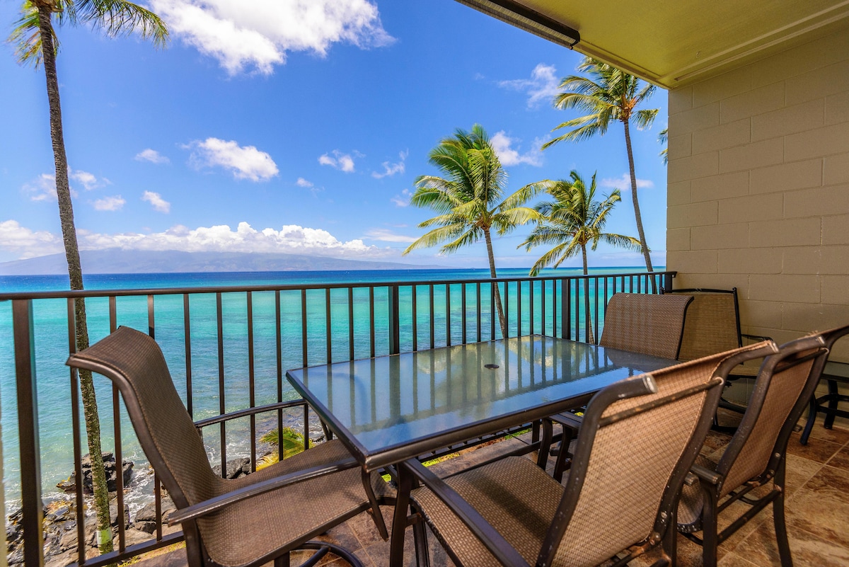A private lanai showcases a glass-top dining table surrounded by comfortable chairs, offering views of the turquoise ocean and palm trees swaying gently in the breeze. The clear blue sky complements the serene beachfront atmosphere.