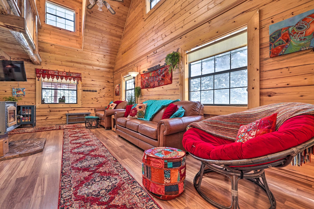 A rustic living area features wooden walls and vaulted ceilings, creating a warm ambiance. A large brown leather sofa is paired with an oversized circular chair, complemented by colorful pillows. A patterned area rug anchors the seating arrangement, while windows allow natural light to illuminate the space.
