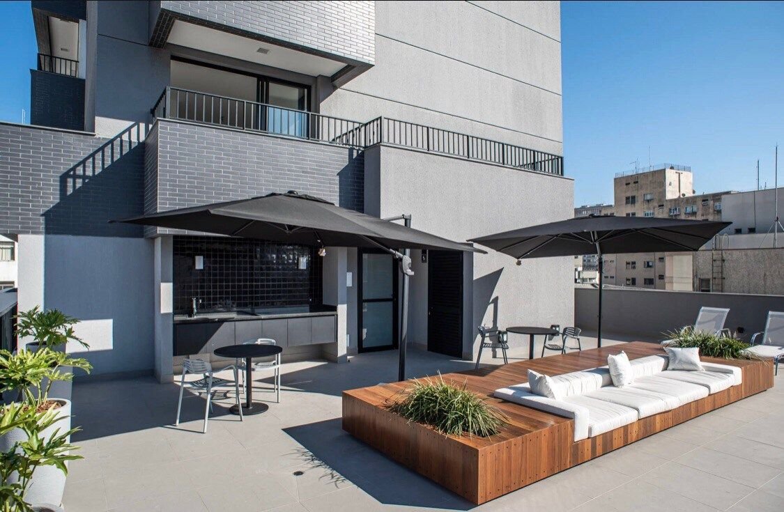 An outdoor terrace features a wooden seating area, complemented by plush cushions and greenery. Two large umbrellas provide shade over a dining space with a round table and chairs. A modern barbecue area is visible, set against a backdrop of the building's architecture.