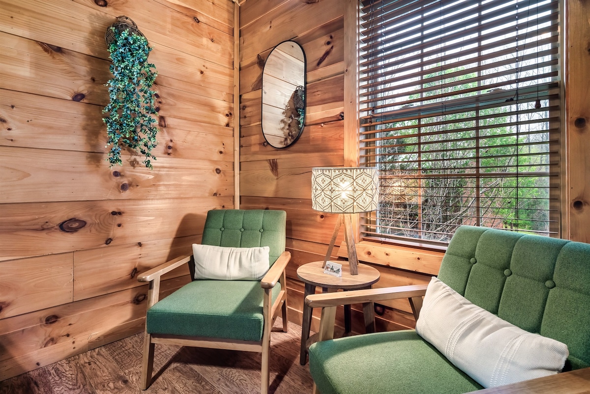 A cozy sitting area features two green armchairs with white cushions, complemented by a small wooden side table. A textured lamp adds soft lighting, while a round mirror reflects the natural wood paneling and sunlight coming through the window, framed by wooden blinds.